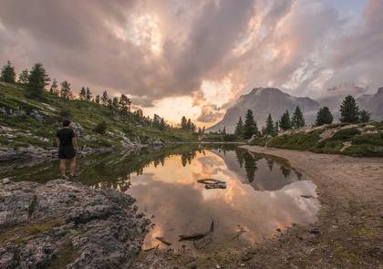A photographer at the edge of an alpine lake at sunset, peaks and cloud reflected in still water.